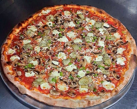 Wood-fired pizza with melted cheese, fresh basil, and assorted toppings on a dark pizza pan